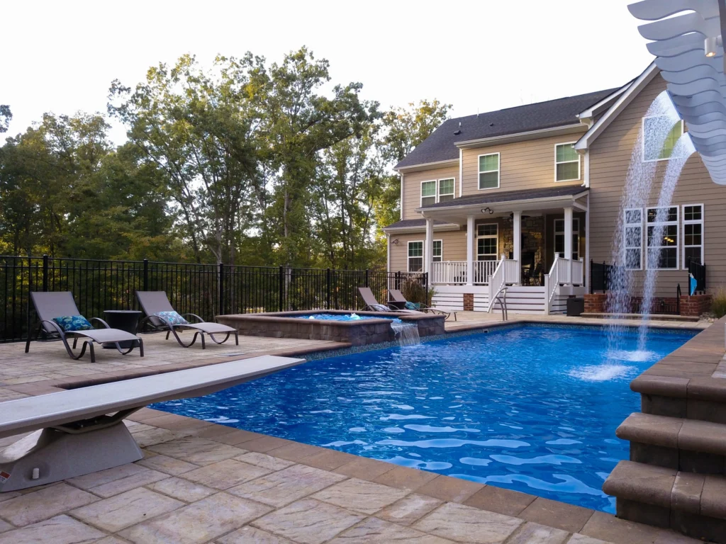 Fiberglass swimming pool with attached spa, surrounded by lounge chairs and a stone patio, featuring a waterfall and residential home backdrop in Bedford, NY.