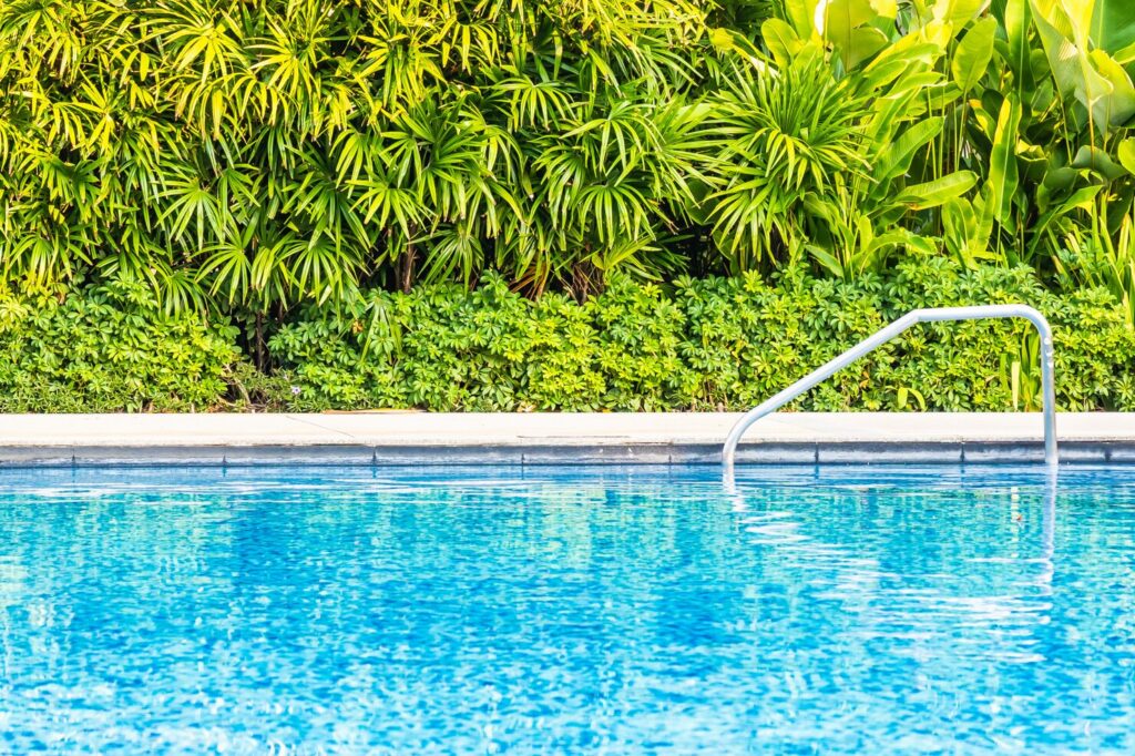 Outdoor fiberglass swimming pool with clear blue water, surrounded by lush green tropical plants and a stainless steel ladder, emphasizing relaxation and vacation vibes.