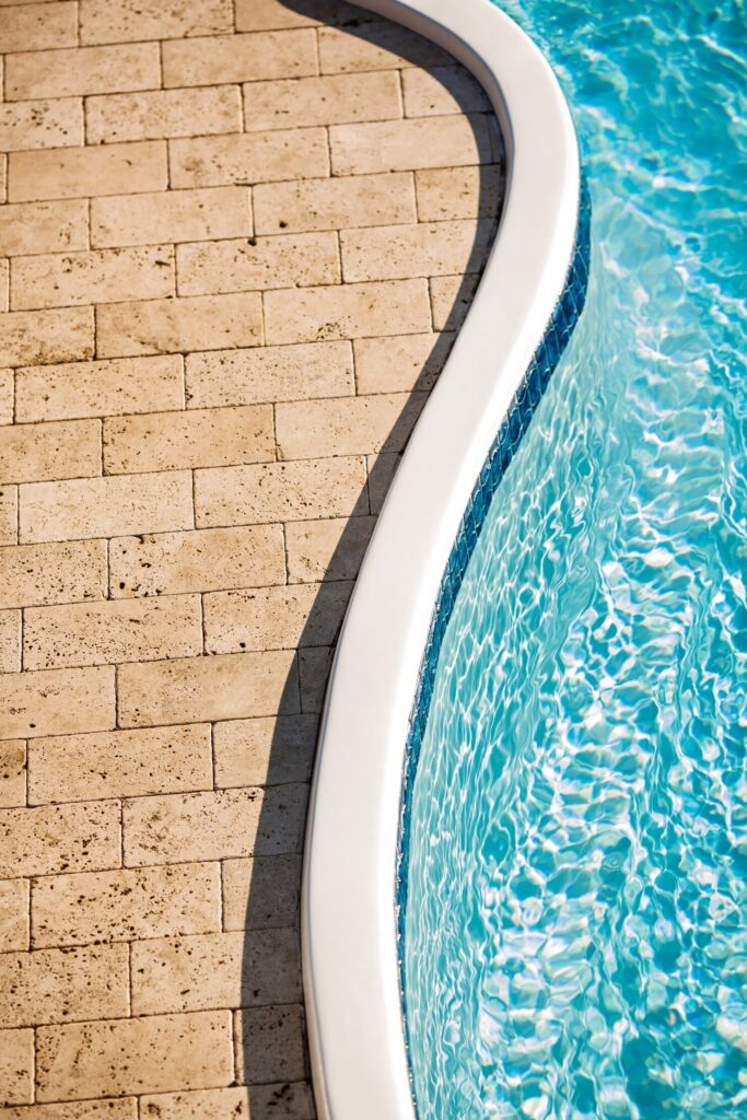 Curved edge of a fiberglass pool with clear blue water and surrounding stone tiles, illustrating design options for West Harrison homes.