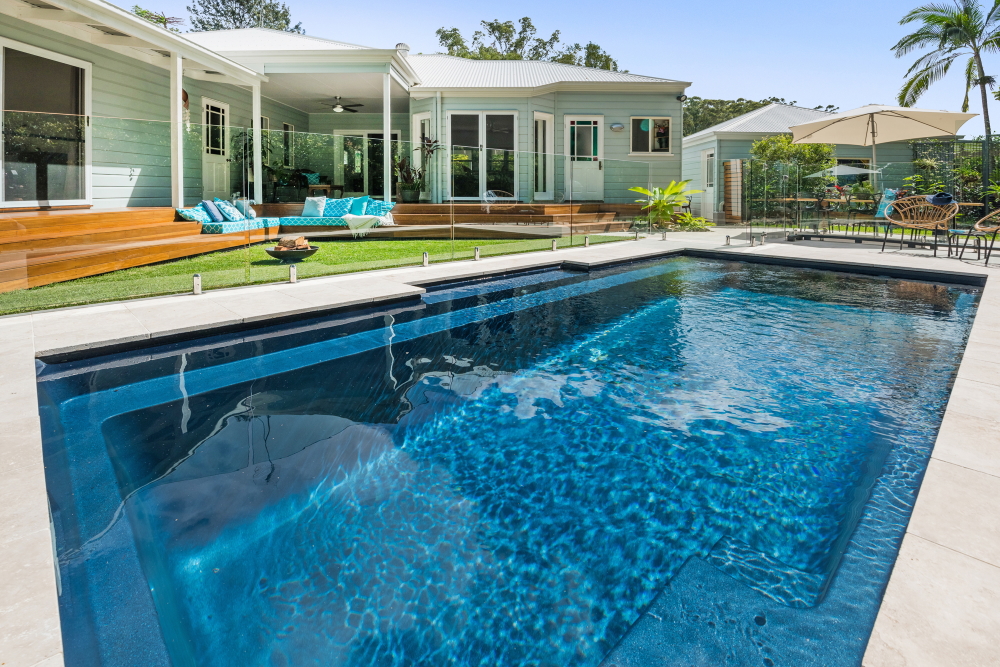 Fiberglass pool with clear blue water, surrounded by a well-designed backyard featuring a wooden deck, lounge seating, and landscaping, highlighting cohesive outdoor living spaces.