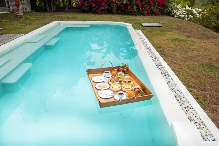 Floating breakfast tray in a swimming pool, featuring fruits, croissants, coffee, and orange juice, surrounded by a tropical resort setting, emphasizing luxury and relaxation for vacationing homeowners.