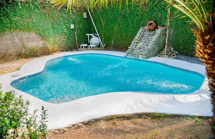 Homemade fiberglass swimming pool surrounded by lush greenery and garden features, showcasing a gentle entrance, filtration equipment, and decorative stone waterfall.