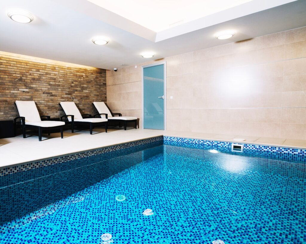 Indoor swimming pool with blue mosaic tiles, surrounded by white lounge chairs and a glass door, emphasizing relaxation and design in home spaces.