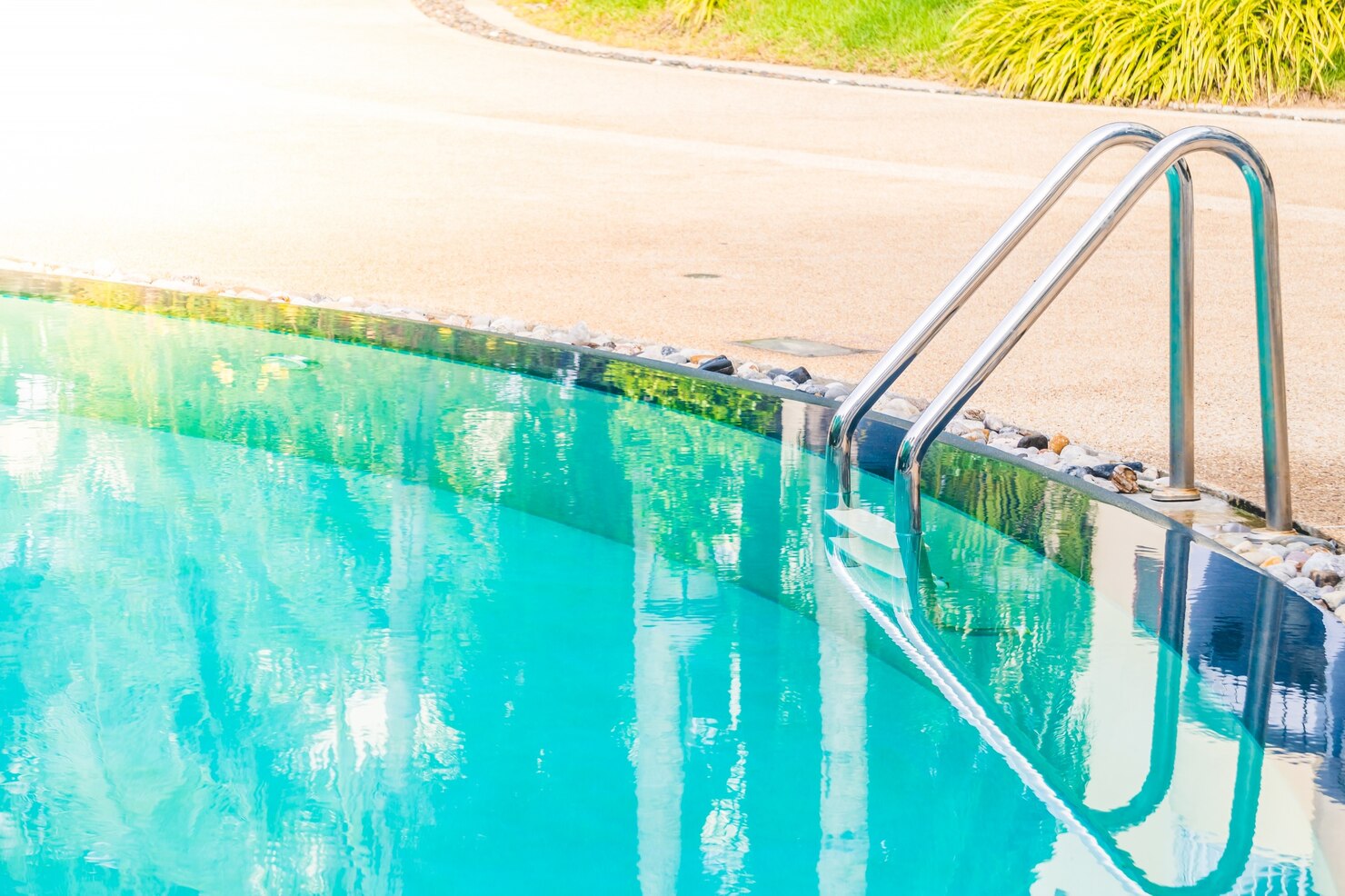 Outdoor fiberglass swimming pool with stainless steel ladder, surrounded by a landscaped yard, reflecting sunlight and greenery.