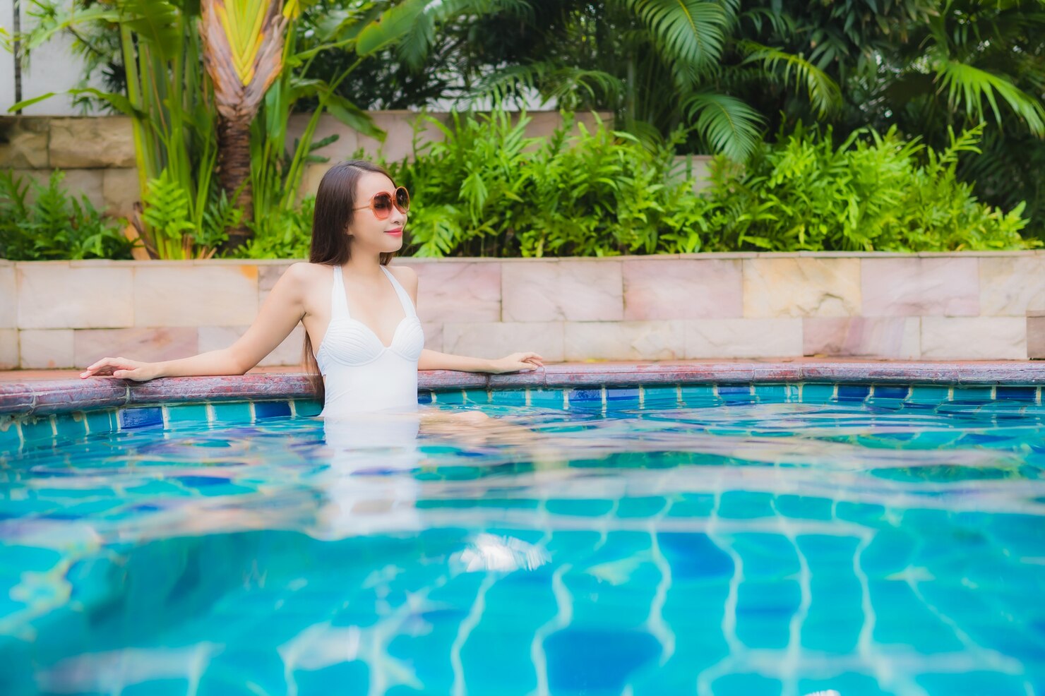 Young woman in white swimsuit relaxing by a fiberglass pool, surrounded by lush greenery, illustrating leisure and poolside enjoyment for Coastal Pools and Spas.