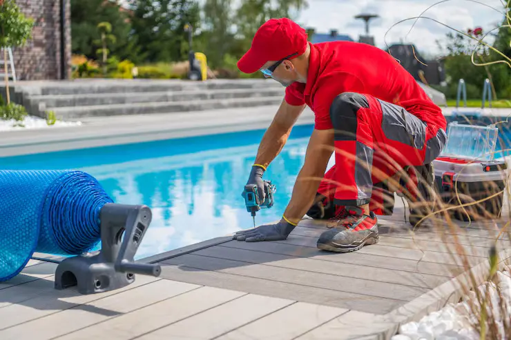Worker installing decking around a fiberglass swimming pool in a residential backyard setting, with pool equipment and landscaping visible.