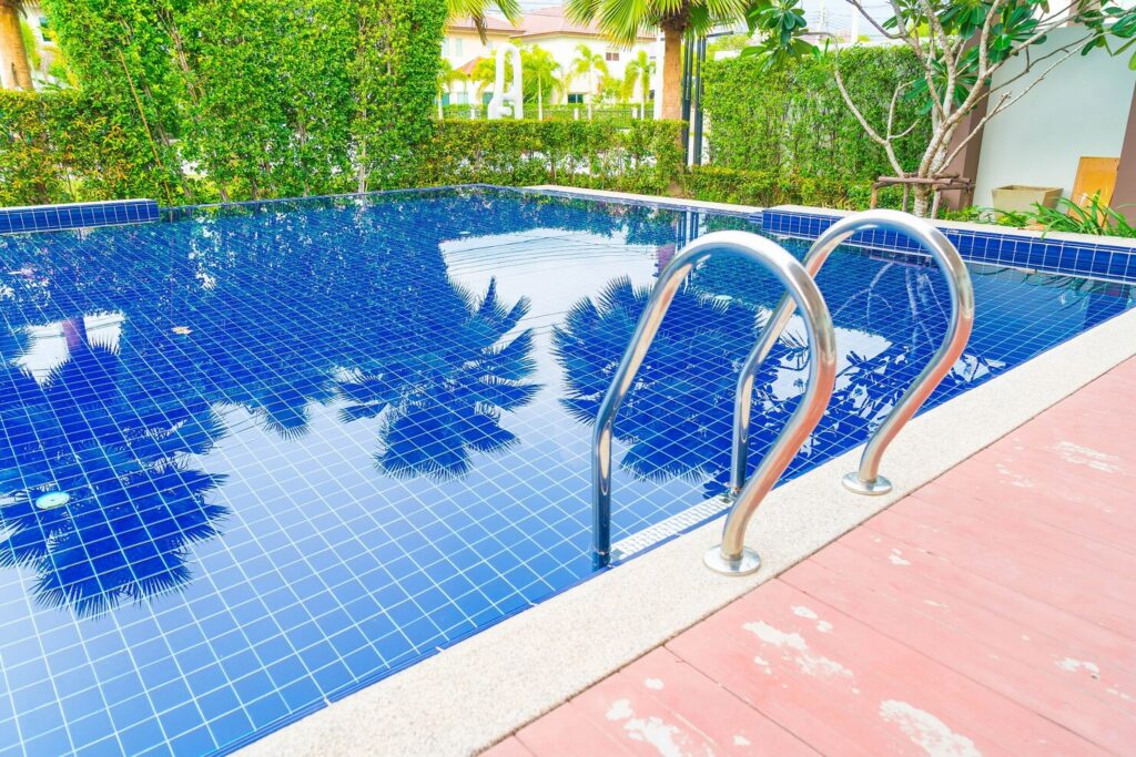Swimming pool with blue tiled surface, stainless steel handrails, surrounded by lush greenery, ideal for backyard designs and family seating areas.
