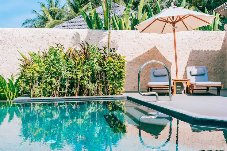 Umbrella and lounge chairs beside a swimming pool with lush greenery and a clear blue sky, highlighting a relaxing outdoor space for fiberglass pool installation.